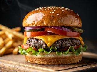 Ein köstlich aussehender Burger mit Zwiebel, Tomate, Gurke, Käse und Salat auf einem Holzbrett vor unscharf erkennbaren Pommes Frites udn einem schwarzem Hintergrund